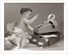 1960s baby seated in small chair hitting keys on office adding machine on top of small file drawers by Anonymous