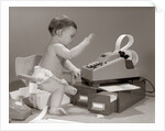 1960s baby seated in small chair hitting keys on office adding machine on top of small file drawers by Anonymous