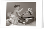 1960s baby seated in small chair hitting keys on office adding machine on top of small file drawers by Anonymous