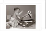 1960s baby seated in small chair hitting keys on office adding machine on top of small file drawers by Anonymous