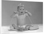 1960s blond baby sitting up looking at camera chewing on telephone cord by Anonymous