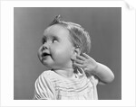 1940s 1950s close-up portrait of baby girl with curl on top of head looking to side with hand held up beside ear by Anonymous