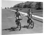1950s teen boy girl couple riding bikes down residential street by Anonymous