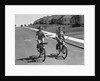 1950s teen boy girl couple riding bikes down residential street by Anonymous