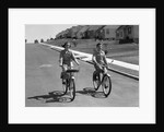1950s teen boy girl couple riding bikes down residential street by Anonymous