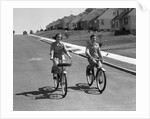 1950s teen boy girl couple riding bikes down residential street by Anonymous