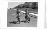 1950s teen boy girl couple riding bikes down residential street by Anonymous