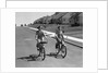 1950s teen boy girl couple riding bikes down residential street by Anonymous