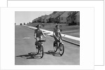 1950s teen boy girl couple riding bikes down residential street by Anonymous