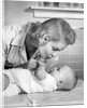 1950s mother woman bending over baby lying on layette holding hands talking by Anonymous