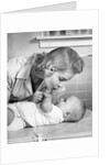 1950s mother woman bending over baby lying on layette holding hands talking by Anonymous