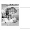 1950s mother woman bending over baby lying on layette holding hands talking by Anonymous