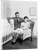 1930s 1940s mother sitting in chair in nursery baby 11 months on lap drinking from bottle father looking on leaning on crib by Anonymous
