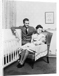 1930s 1940s mother sitting in chair in nursery baby 11 months on lap drinking from bottle father looking on leaning on crib by Anonymous