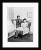 1930s 1940s mother sitting in chair in nursery baby 11 months on lap drinking from bottle father looking on leaning on crib by Anonymous