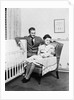 1930s 1940s mother sitting in chair in nursery baby 11 months on lap drinking from bottle father looking on leaning on crib by Anonymous