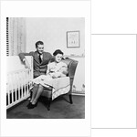 1930s 1940s mother sitting in chair in nursery baby 11 months on lap drinking from bottle father looking on leaning on crib by Anonymous