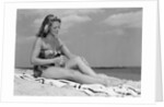 1950s woman in strapless one-piece bathing suit seated on beach towel putting on suntan lotion by Corbis