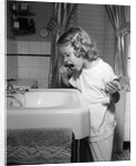 1950s little blond girl in pajamas standing at bathroom sink brushing her teeth toothpaste and toothbrush in hand by Anonymous