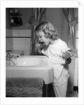 1950s little blond girl in pajamas standing at bathroom sink brushing her teeth toothpaste and toothbrush in hand by Anonymous