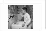 1950s boy combing hair looking in bathroom sink mirror by Anonymous