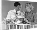1950s smiling parents picking baby up out of crib by Anonymous