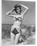 1950s smiling young woman kneeling in grassy sand wearing polka dot bikini shading eyes from sun by Anonymous