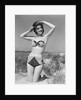 1950s smiling young woman kneeling in grassy sand wearing polka dot bikini shading eyes from sun by Anonymous