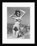 1950s smiling young woman kneeling in grassy sand wearing polka dot bikini shading eyes from sun by Anonymous