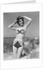 1950s smiling young woman kneeling in grassy sand wearing polka dot bikini shading eyes from sun by Anonymous