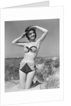1950s smiling young woman kneeling in grassy sand wearing polka dot bikini shading eyes from sun by Anonymous
