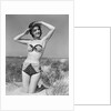 1950s smiling young woman kneeling in grassy sand wearing polka dot bikini shading eyes from sun by Anonymous
