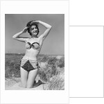 1950s smiling young woman kneeling in grassy sand wearing polka dot bikini shading eyes from sun by Anonymous