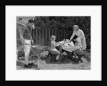 1960s man on patio grilling steak with 2 daughters seated at picnic table & wife standing serving food by Anonymous