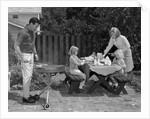 1960s man on patio grilling steak with 2 daughters seated at picnic table & wife standing serving food by Anonymous