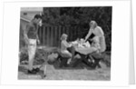 1960s man on patio grilling steak with 2 daughters seated at picnic table & wife standing serving food by Anonymous
