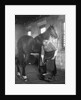 1930s elderly bearded blacksmith shoeing horse holding hoof between legs & hammer in other hand by Anonymous