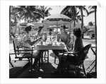 1930s three women and one man sitting at tropical pool side table talking together by Anonymous