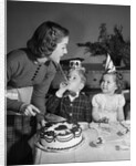 1950s woman mother cutting birthday cake for two children sitting at table wearing party hats by Anonymous