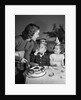 1950s woman mother cutting birthday cake for two children sitting at table wearing party hats by Anonymous