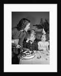 1950s woman mother cutting birthday cake for two children sitting at table wearing party hats by Anonymous
