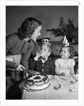 1950s woman mother cutting birthday cake for two children sitting at table wearing party hats by Anonymous