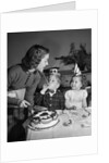 1950s woman mother cutting birthday cake for two children sitting at table wearing party hats by Anonymous