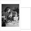1950s woman mother cutting birthday cake for two children sitting at table wearing party hats by Anonymous
