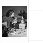 1950s woman mother cutting birthday cake for two children sitting at table wearing party hats by Anonymous