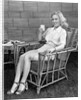 1940s blonde woman in bamboo chair holding up glass of milk smiling wearing white shorts looking at camera by Anonymous