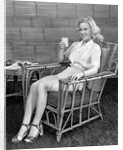 1940s blonde woman in bamboo chair holding up glass of milk smiling wearing white shorts looking at camera by Anonymous