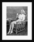 1940s blonde woman in bamboo chair holding up glass of milk smiling wearing white shorts looking at camera by Anonymous