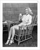 1940s blonde woman in bamboo chair holding up glass of milk smiling wearing white shorts looking at camera by Anonymous
