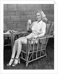 1940s blonde woman in bamboo chair holding up glass of milk smiling wearing white shorts looking at camera by Anonymous